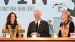John Waters, Aubrey Plaza, and Amy Sedaris appearing together on “The View.” Three hosts seated behind a wooden desk on a TV talk show set, with a large gold 'THE VIEW' logo in front of the left host and coffee mugs.