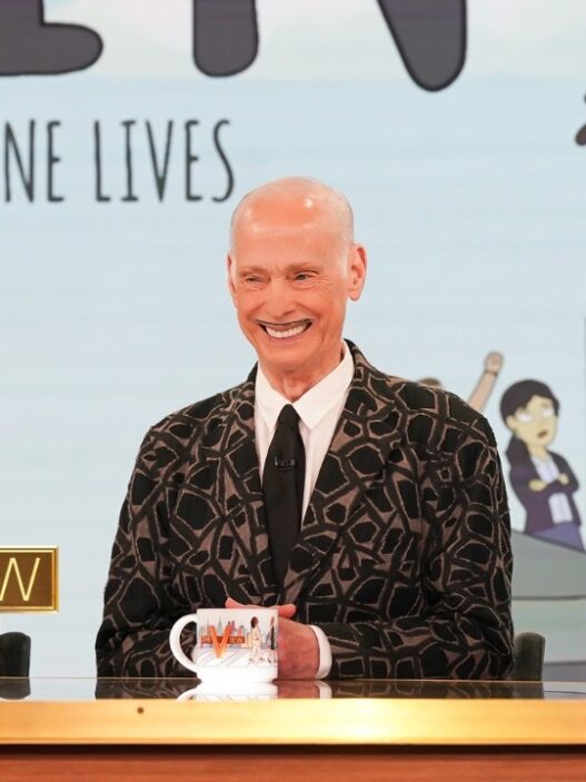 John Waters, Aubrey Plaza, and Amy Sedaris appearing together on “The View.” Three hosts seated behind a wooden desk on a TV talk show set, with a large gold 'THE VIEW' logo in front of the left host and coffee mugs.