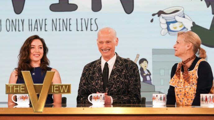 John Waters, Aubrey Plaza, and Amy Sedaris appearing together on “The View.” Three hosts seated behind a wooden desk on a TV talk show set, with a large gold 'THE VIEW' logo in front of the left host and coffee mugs.