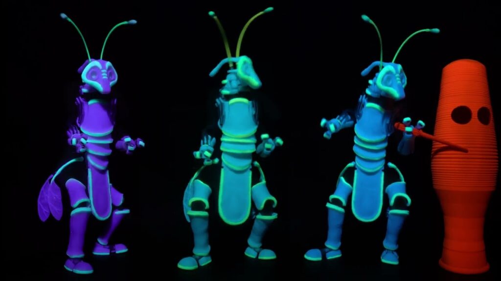 Three neon-lit insect puppets with antennae pose on a dark stage under blacklight, beside an orange pin-shaped prop.