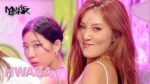 Two female performers in a pink-lit studio, the woman with red hair smiling toward the camera; Music Bank logo in the corner.