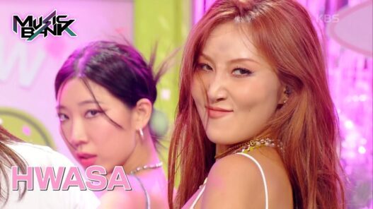 Two female performers in a pink-lit studio, the woman with red hair smiling toward the camera; Music Bank logo in the corner.