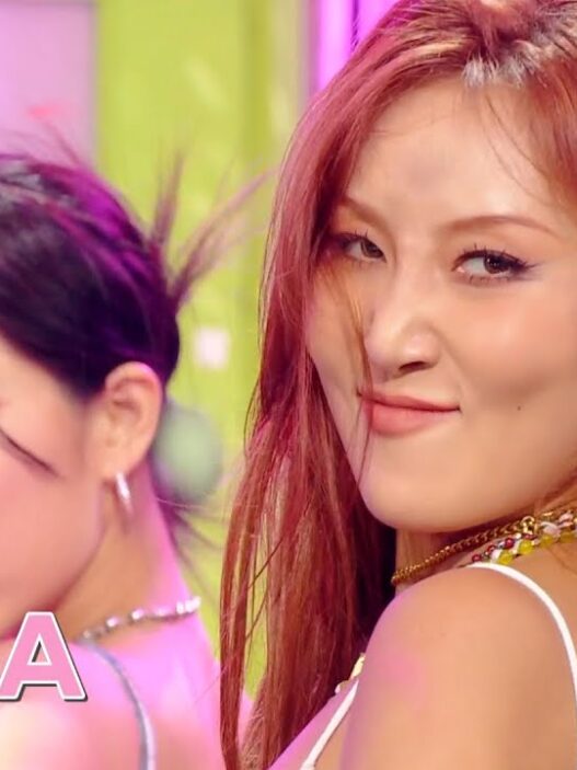 Two female performers in a pink-lit studio, the woman with red hair smiling toward the camera; Music Bank logo in the corner.