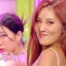 Two female performers in a pink-lit studio, the woman with red hair smiling toward the camera; Music Bank logo in the corner.