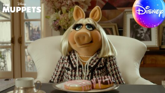 Miss Piggy sits at a desk wearing a tweed jacket and pearls, with two donuts on a plate in front of her; Disney Muppets promo scene.