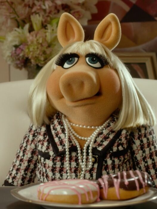 Miss Piggy sits at a desk wearing a tweed jacket and pearls, with two donuts on a plate in front of her; Disney Muppets promo scene.