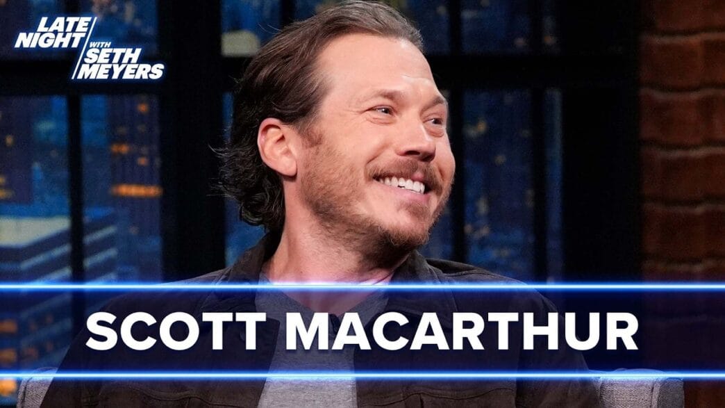 Scott Macarthur smiles on Late Night with Seth Meyers set, with a blue banner displaying his name across the bottom, studio lights in the background.
