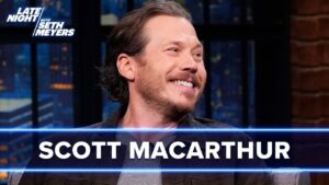 Scott Macarthur smiles on Late Night with Seth Meyers set, with a blue banner displaying his name across the bottom, studio lights in the background.