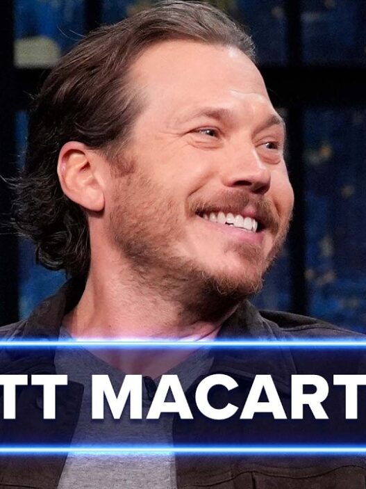Scott Macarthur smiles on Late Night with Seth Meyers set, with a blue banner displaying his name across the bottom, studio lights in the background.