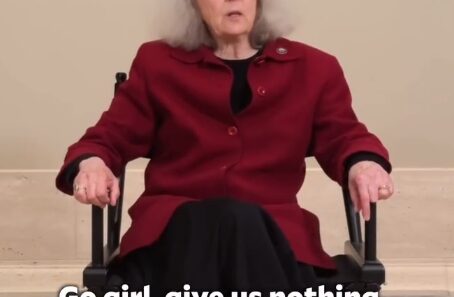 Older woman in a red coat seated in a wheelchair, speaking to the camera; caption in image reads: “Go girl, give us nothing.” indoors in a neutral setting.