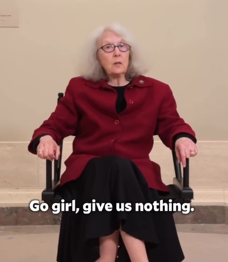Older woman in a red coat seated in a wheelchair, speaking to the camera; caption in image reads: “Go girl, give us nothing.” indoors in a neutral setting.