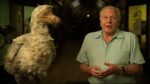 Older man in a pale green shirt stands beside a large, fluffy pale bird (likely taxidermy) in a museum.