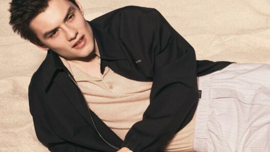 Young man lying on sandy beach, wearing a beige shirt and black jacket, looking to the side in a relaxed pose.