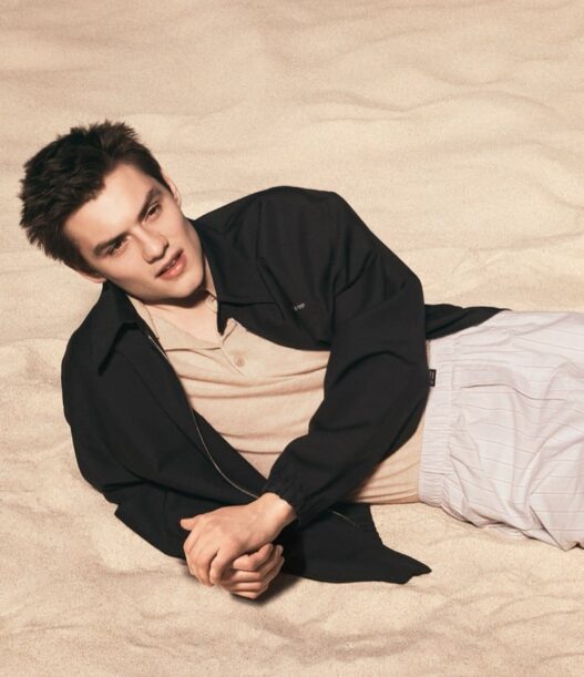 Young man lying on sandy beach, wearing a beige shirt and black jacket, looking to the side in a relaxed pose.