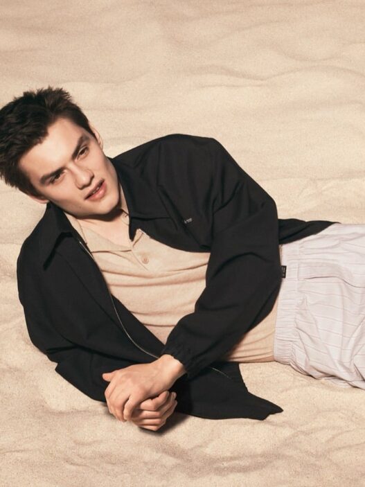 Young man lying on sandy beach, wearing a beige shirt and black jacket, looking to the side in a relaxed pose.