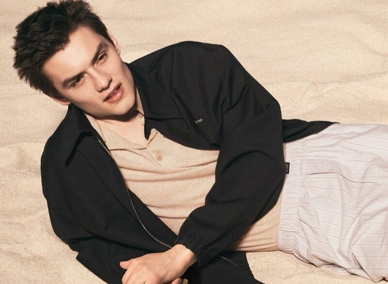 Young man lying on sandy beach, wearing a beige shirt and black jacket, looking to the side in a relaxed pose.