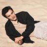 Young man lying on sandy beach, wearing a beige shirt and black jacket, looking to the side in a relaxed pose.