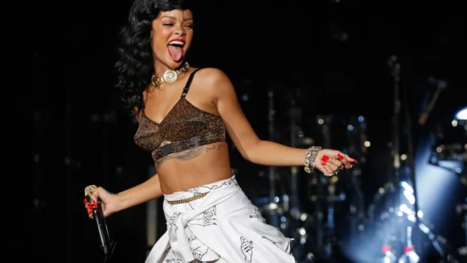 Rihanna-style singer performing on stage in a leopard-print crop top, white patterned skirt, and gold jewelry, microphone in hand with tongue out.