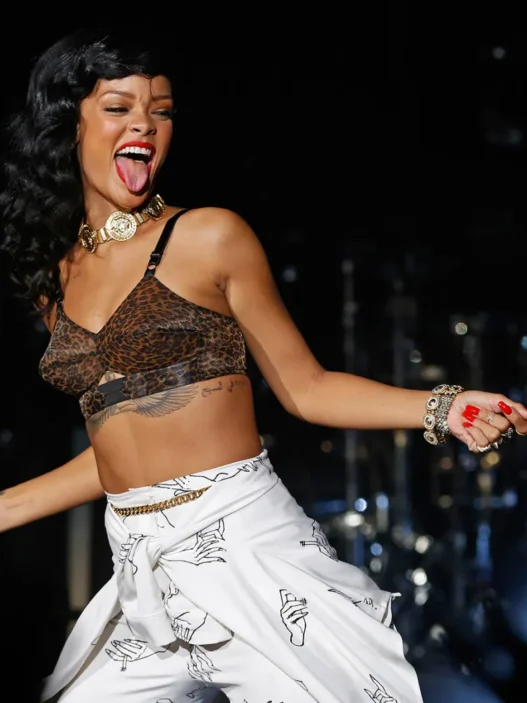 Rihanna-style singer performing on stage in a leopard-print crop top, white patterned skirt, and gold jewelry, microphone in hand with tongue out.