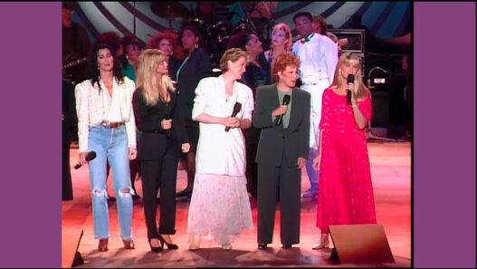 Group of singers on stage performing into handheld microphones, with backup singers behind them.