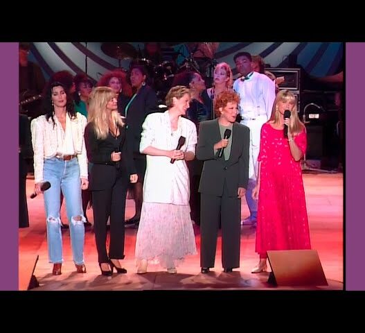 Group of singers on stage performing into handheld microphones, with backup singers behind them.