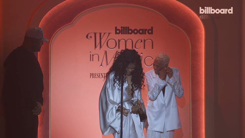 Two women in white outfits smiling on a pink stage at Billboard's Women in Music event, with the sign Reading 'billboard Women in Music' behind them, a microphone in front.