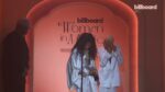 Two women in white outfits smiling on a pink stage at Billboard's Women in Music event, with the sign Reading 'billboard Women in Music' behind them, a microphone in front.