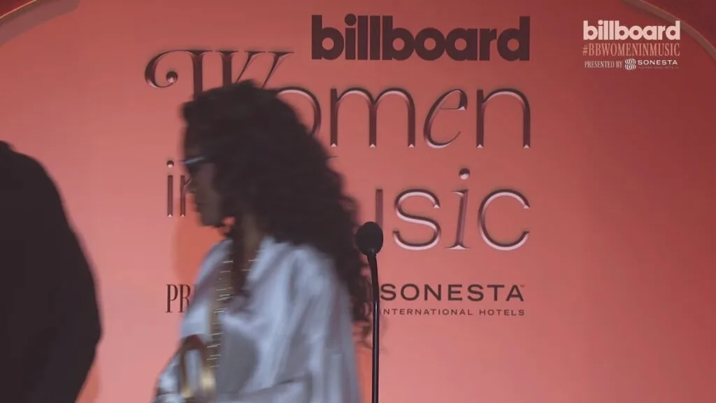 Blurred woman in a white shirt walking past a pink Billboard Women in Music backdrop with a microphone in front.