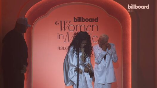 Two women in white outfits smiling on a pink stage at Billboard's Women in Music event, with the sign Reading 'billboard Women in Music' behind them, a microphone in front.