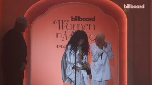 Two women in white outfits smiling on a pink stage at Billboard's Women in Music event, with the sign Reading 'billboard Women in Music' behind them, a microphone in front.