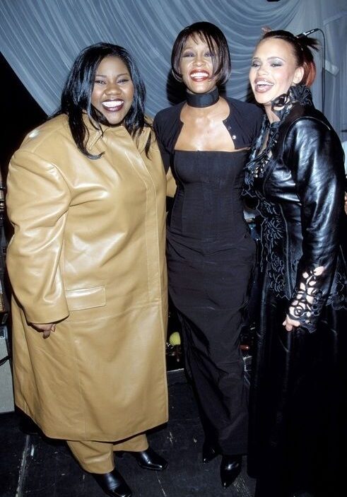 Three women posing together and smiling at a backstage event, wearing stylish outfits: camel coat, black jumpsuit, and black leather coat with sheer scarf detail.",