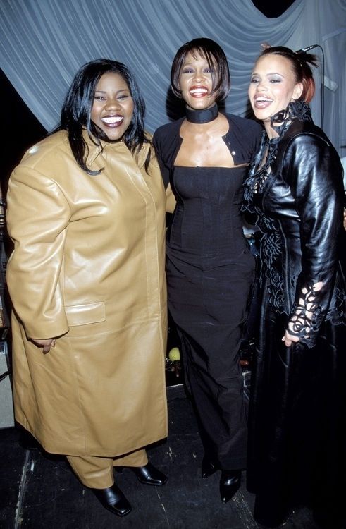 Three women posing together and smiling at a backstage event, wearing stylish outfits: camel coat, black jumpsuit, and black leather coat with sheer scarf detail.",