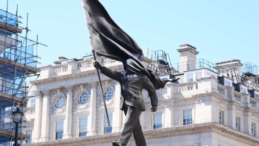 Bronze statue of a man in a suit with a billowing cape, walking forward on a pedestal in an urban square against a historic building backdrop.