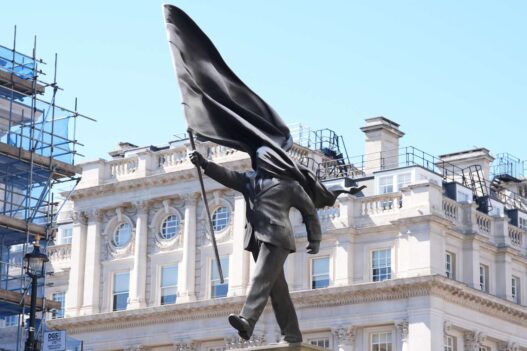 Bronze statue of a man in a suit with a billowing cape, walking forward on a pedestal in an urban square against a historic building backdrop.