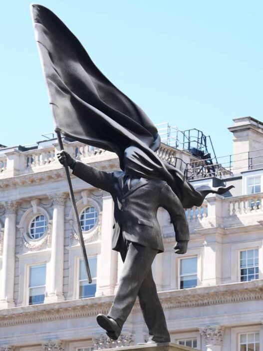 Bronze statue of a man in a suit with a billowing cape, walking forward on a pedestal in an urban square against a historic building backdrop.