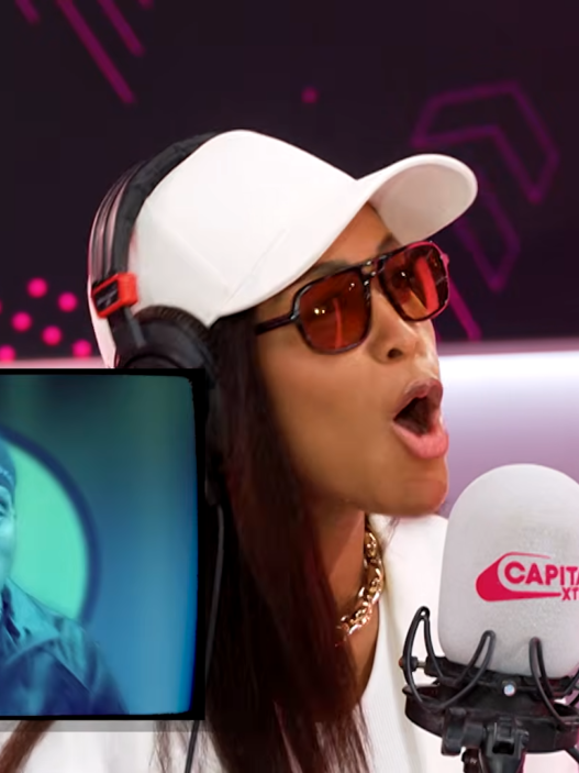 Radio host wearing white cap and red sunglasses speaks into a Capital Xtra microphone while a blue-tinted screen shows a guest.