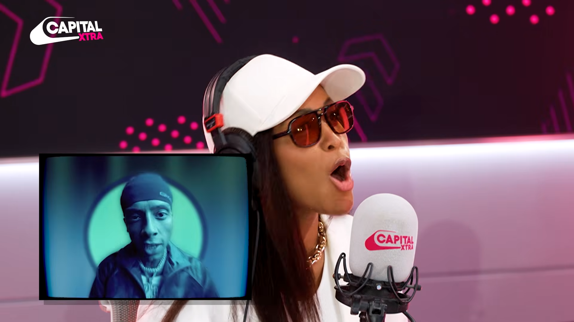 Radio host wearing white cap and red sunglasses speaks into a Capital Xtra microphone while a blue-tinted screen shows a guest.