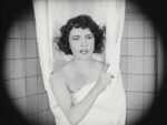 A black-and-white photo of a woman wrapped in a towel, standing between shower curtains, gesturing with her finger as she speaks.