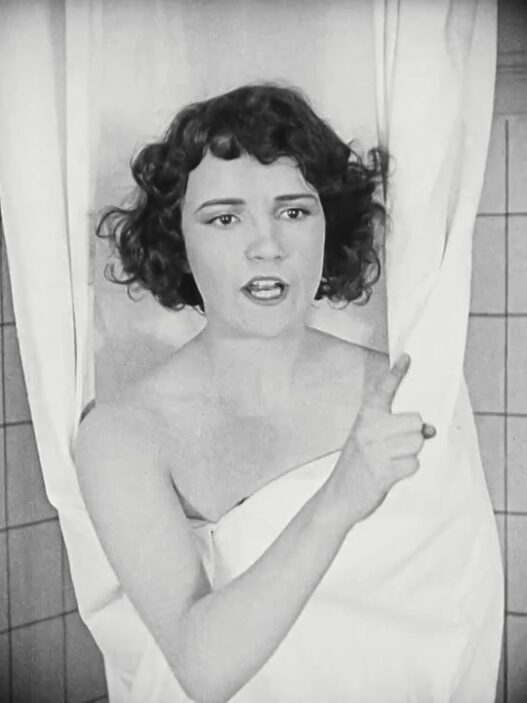A black-and-white photo of a woman wrapped in a towel, standing between shower curtains, gesturing with her finger as she speaks.