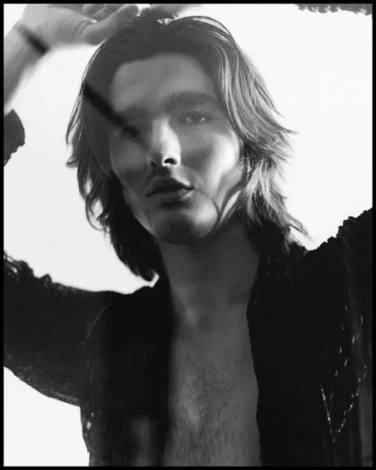 Black-and-white portrait of a young man with shoulder-length hair, shirt open, posed with hands near head.