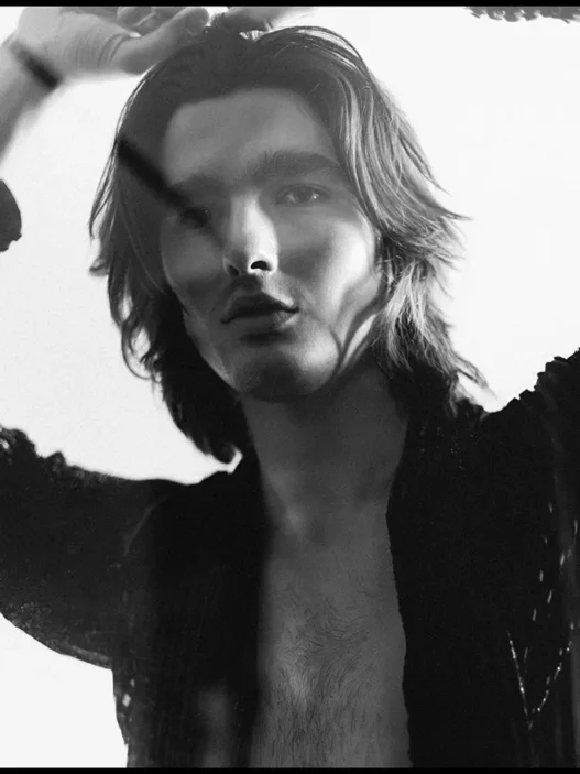 Black-and-white portrait of a young man with shoulder-length hair, shirt open, posed with hands near head.