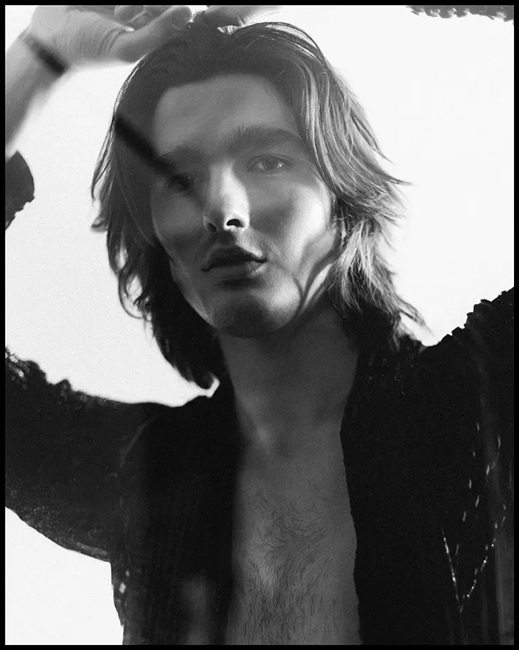 Black-and-white portrait of a young man with shoulder-length hair, shirt open, posed with hands near head.