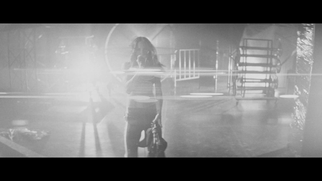 Silhouetted person in a dim studio holding a guitar, with stacked chairs and reflective floor in the background (grainy black-and-white image).