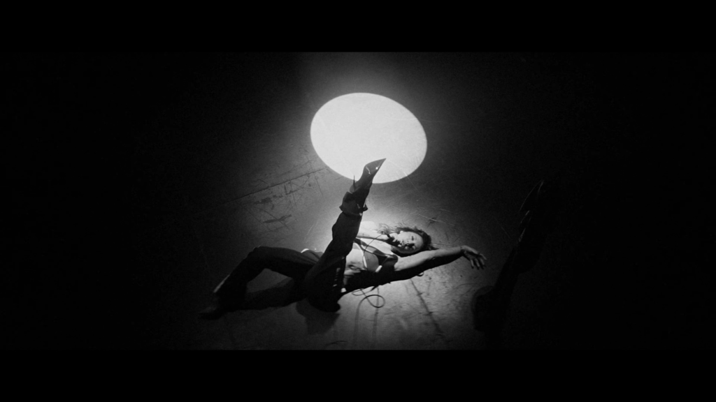 Black-and-white photo of a performer lying on the floor, reaching toward a large glowing circle in a dark, abstract stage setting.