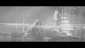 Person lying on the floor with arms outstretched in a dim indoor space; a blurred figure stands in the background.