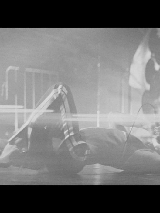 Person lying on the floor with arms outstretched in a dim indoor space; a blurred figure stands in the background.