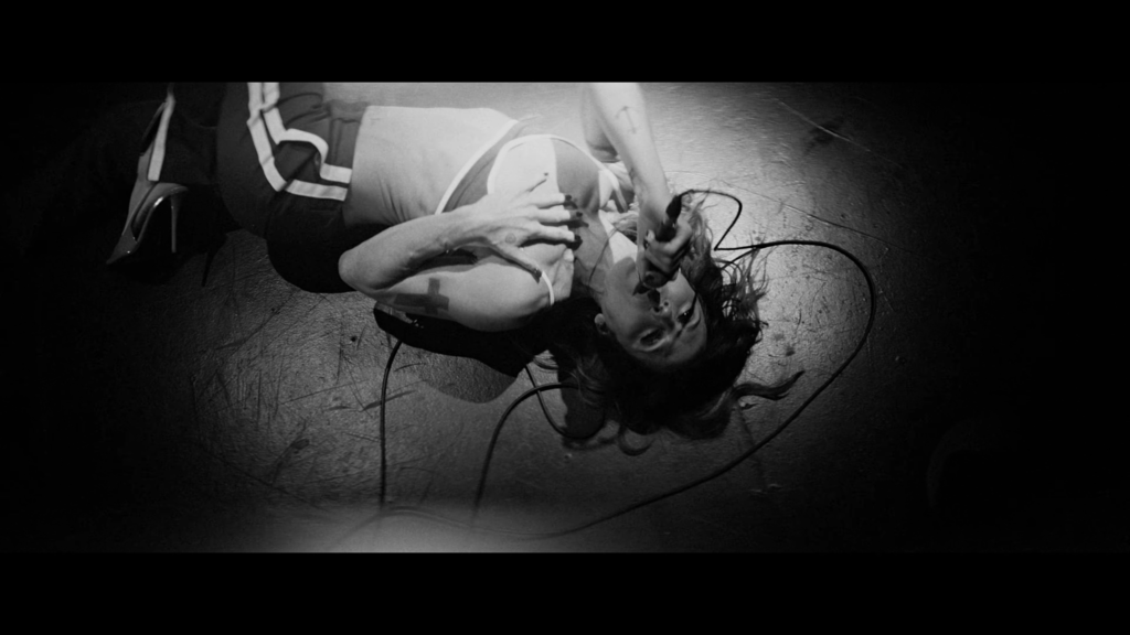 Black-and-white photo of a woman lying on a stage floor, tangled with cables, hair spread around, wearing a sleeveless top.