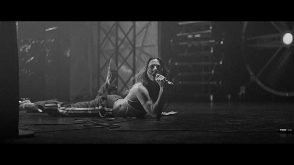 Black-and-white photo of a female singer lying on her stomach on stage, singing into a handheld microphone.