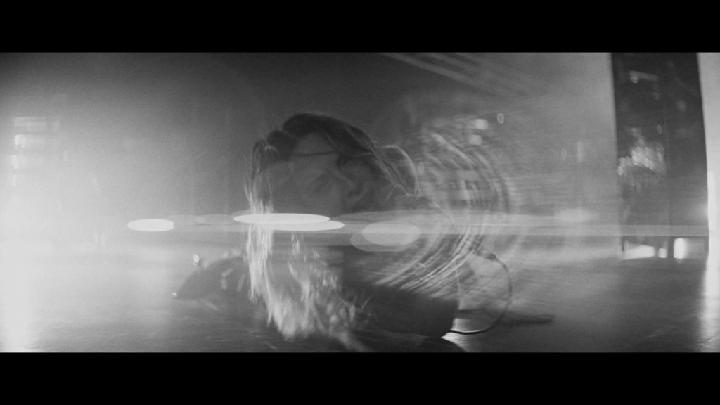 Abstract black-and-white portrait: person with long hair, seen as a blurred reflection with circular light bokeh across a glass surface.