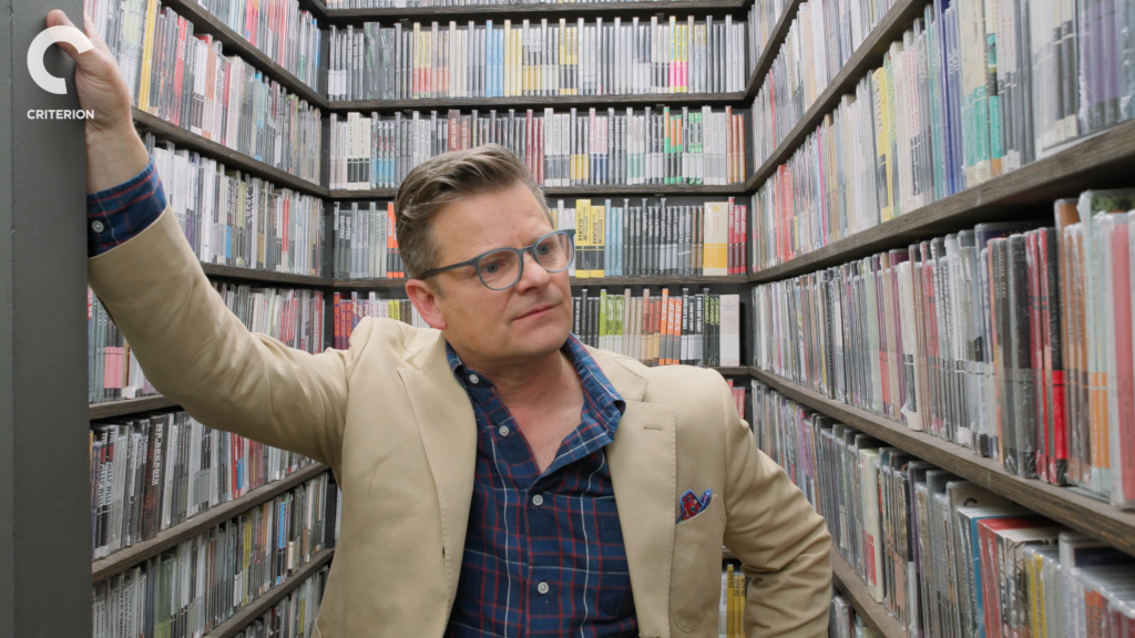 Man wearing glasses and a beige blazer leans against a pillar in a narrow aisle lined with shelves of VHS tapes.
